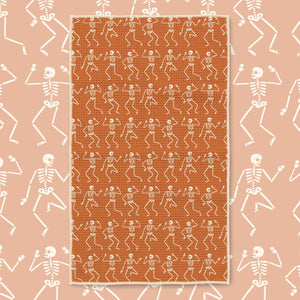 An orange rectangular rug with a repeating pattern of white dancing skeletons, centered on a peach background with a matching skeleton border.